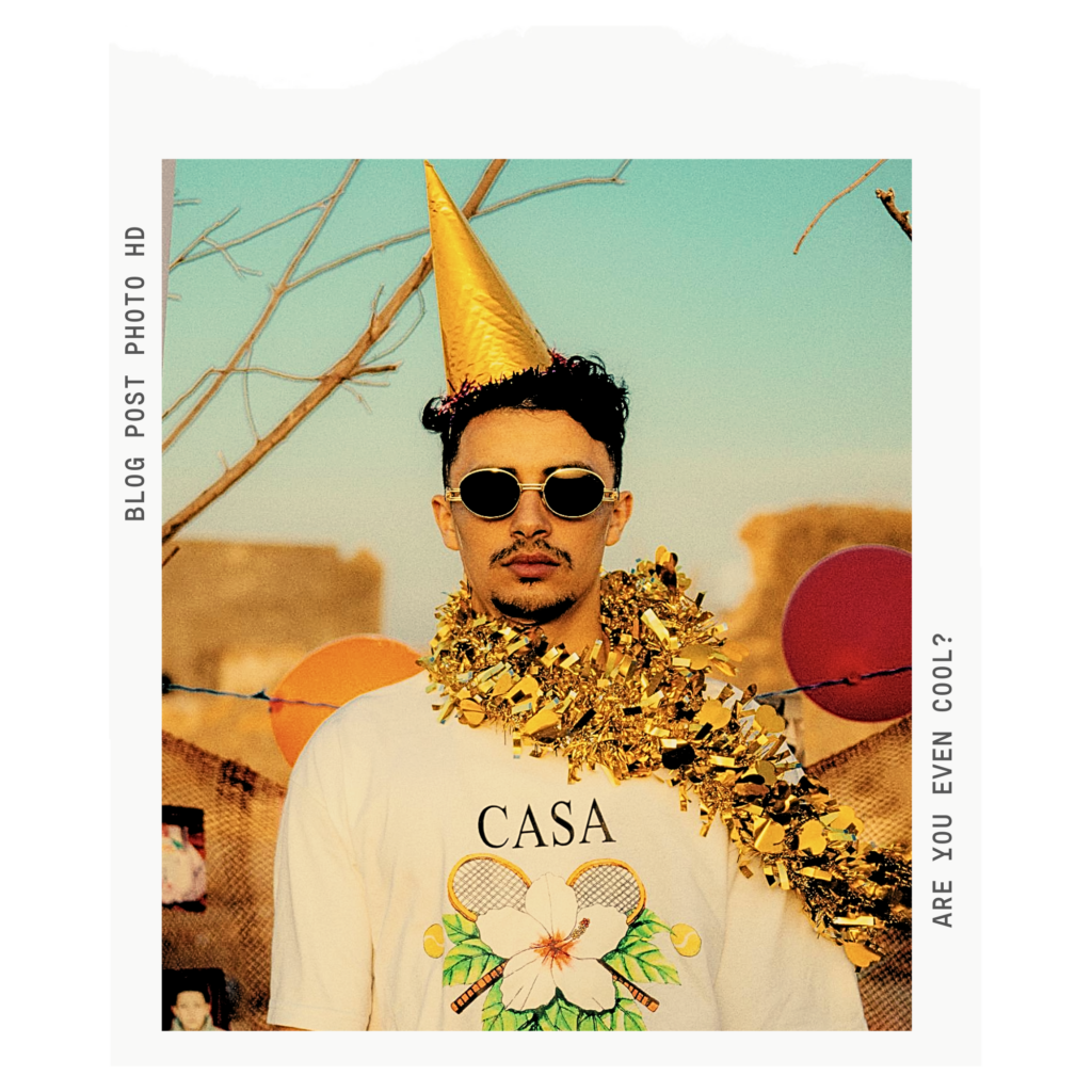 man looking uncelebratory while wearing a confetti boa and a party hat and sunglasses like he's too cool for the party